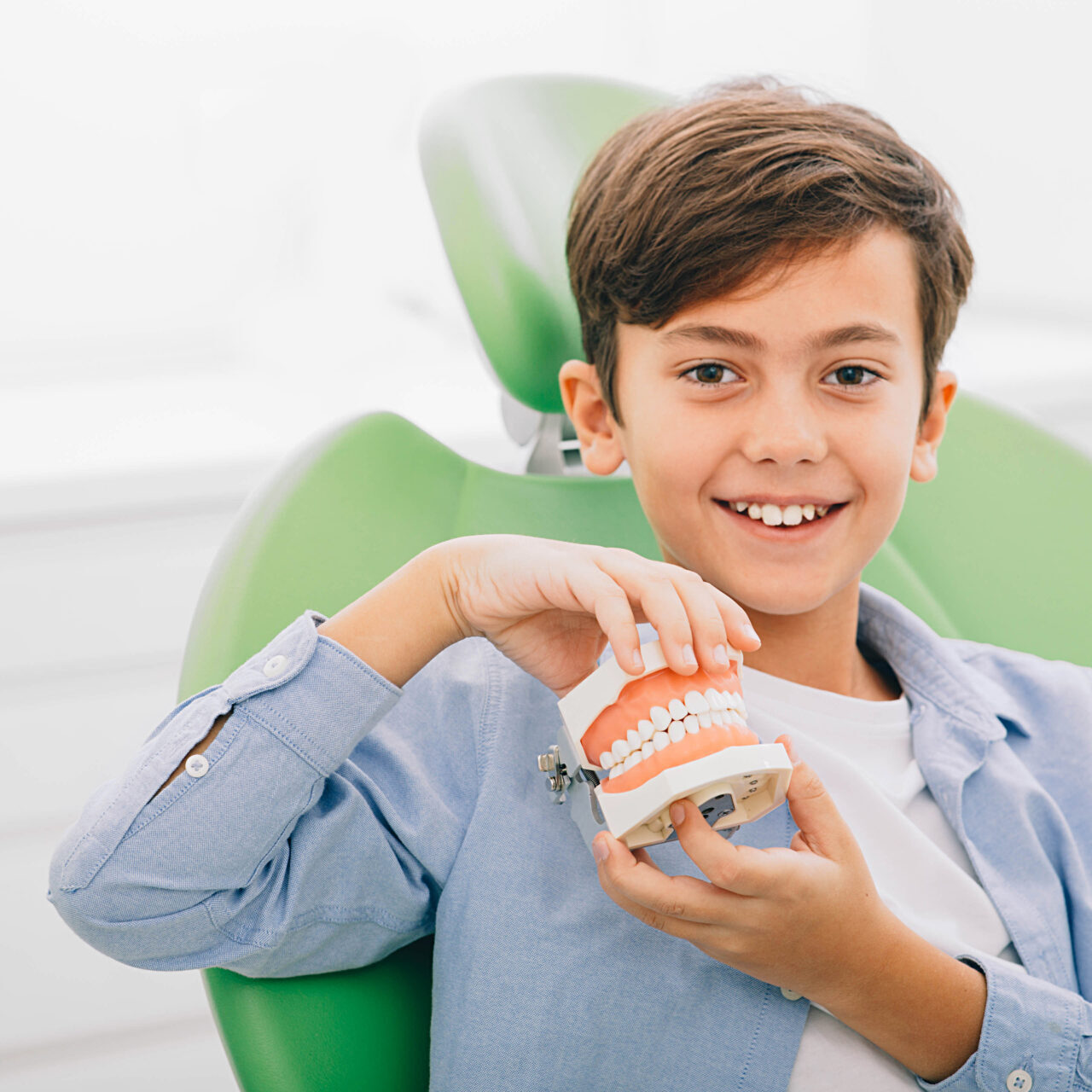 Mixed-race boy holding dental jaw while sitting in dental chair. He is happy after teeth treatment